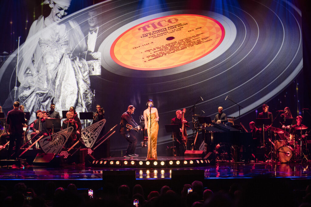 A woman in a long strapless gold gown sings on stage in front of a symphony; in the background is a projection of a record called TICO by Tito Puente and a black and white photo of a woman.