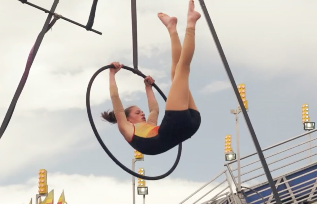 A woman wearing a leotard holds onto the top of a suspended hoop and swings her legs up.