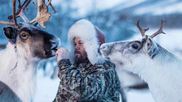A man wearing a fur-lined hooded coat feeds a reindeer by hand while another waits their turn.