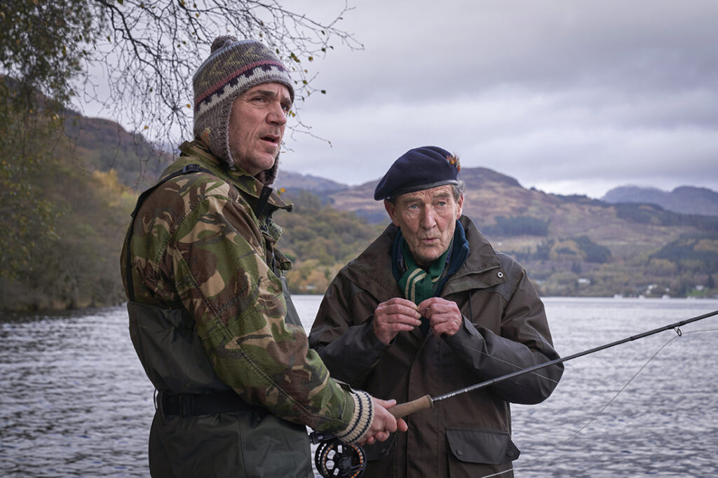 Two men stand in front of a lake and hills, one holds a fishing pole, both wearing heavy coats and hats.