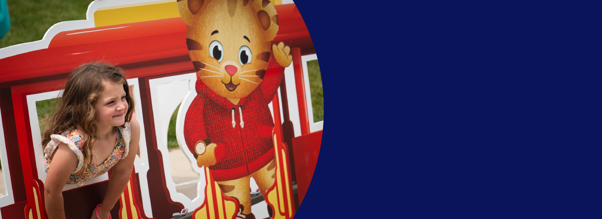 A young girl leans out through the window of a cardboard red trolley car with a tiger character wearing a red sweater standing in front of the trolley car.