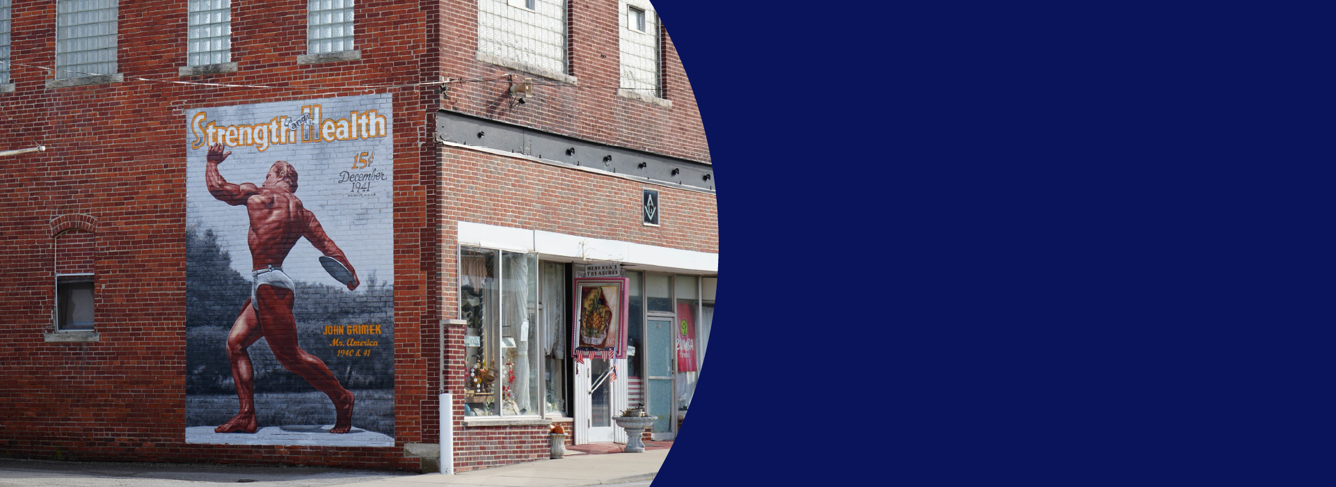 An old brick building with a mural on the side showing a vintage magazine cover called "Strength and Health" with a muscular man holding a discus and wearing white briefs. Next to him is printed "John Grimek, Mr. America 1940 & 41". On the front of the building are storefronts for a gift shop and more.