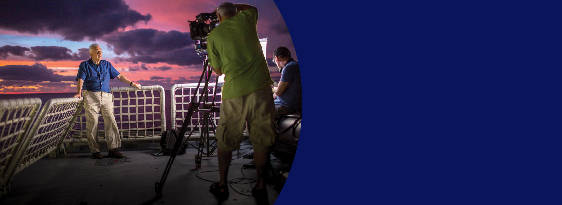 A TV crew films an older man wearing a blue button-down shirt and khaki pants. He leans against white divides, a beautiful pink, purple and blue sunset is in the background.