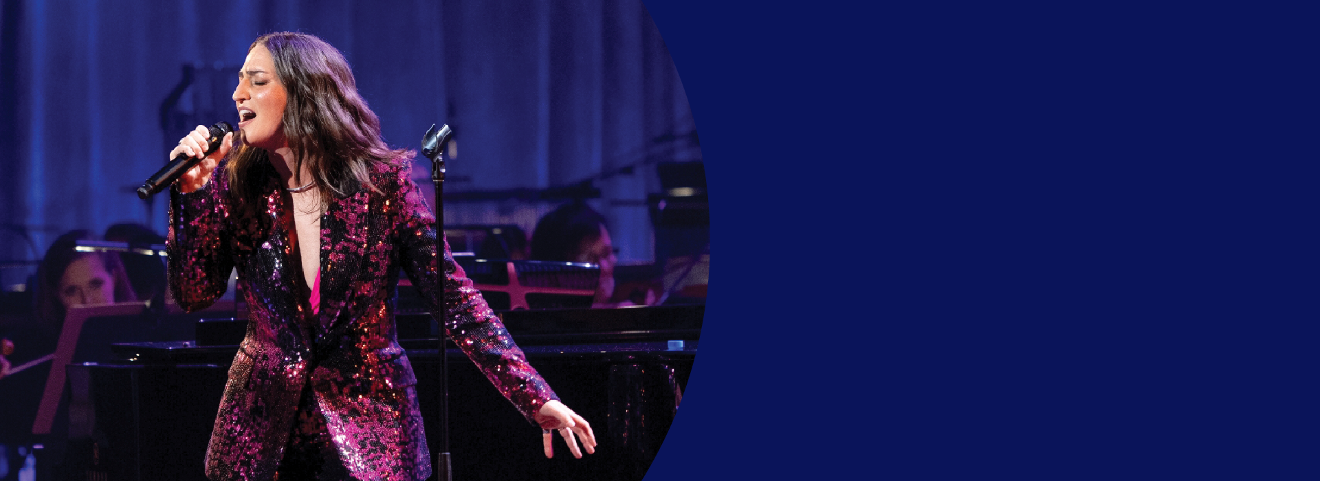 A woman wearing a purple sequined suit sings into a microphone with an orchestra behind her