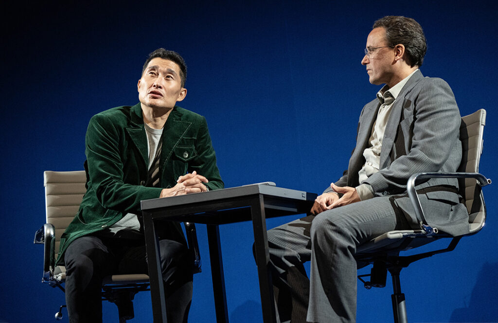 Two men sit across from one another at a small table sitting on desk chairs. The man on the left is wearing a velvet green suit jacket and looking up towards the the stage lights. The man on the right wears an all gray suit and looks at the other man. Both men sit in the chairs with their hands clasped together.