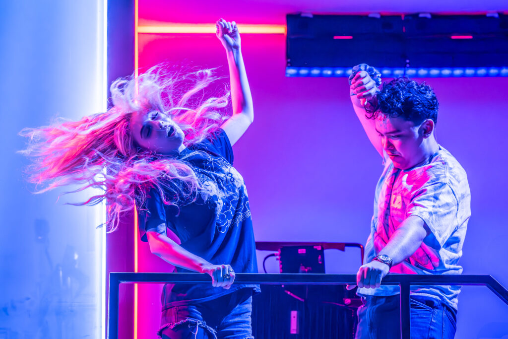 Two people are dancing energetically under vibrant neon lighting in shades of blue and pink. The woman has long, flowing hair and is wearing a graphic T-shirt and shorts, while the man wears a tie-dye shirt and jeans, both immersed in the moment.