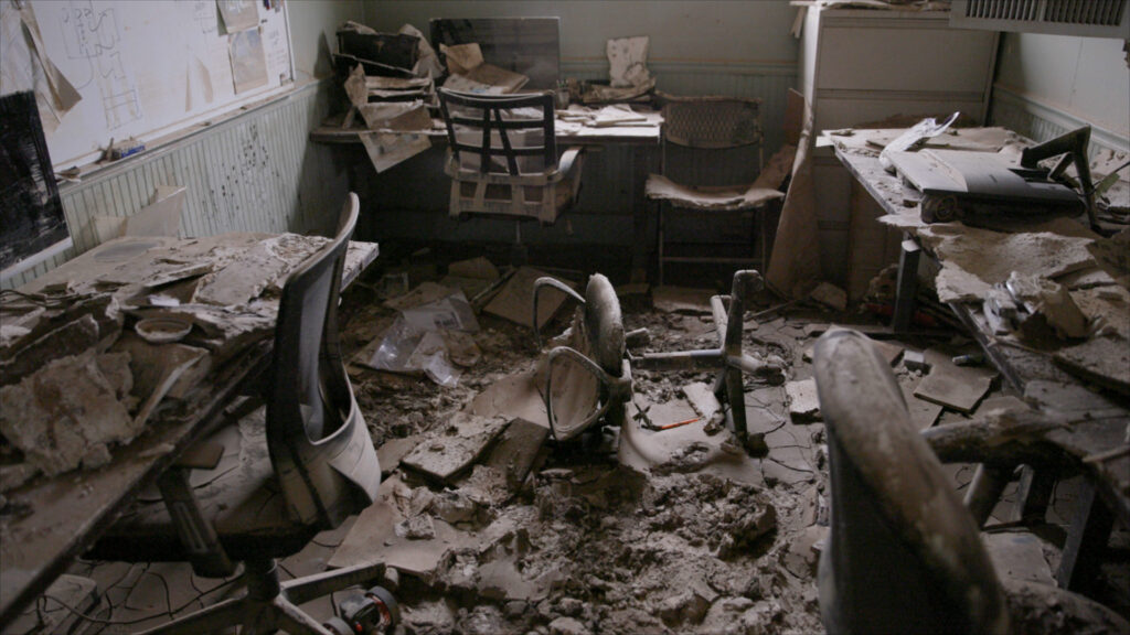 The image shows a severely damaged and abandoned office room, the aftermath of the hurricane. The room is in chaos: Debris covers the floor, including broken ceiling tiles, plaster, and dirt. Several office chairs are overturned or broken, with one in the center lying on its side, caked in mud. Desks are cluttered with water-damaged papers, crumbling drywall, and broken office equipment. One desk has what appears to be a soaked and unusable printer or scanner. The walls and floor are stained and cracked, with mold and mud streaks adding to the atmosphere of decay. A whiteboard on the left wall still has faded markings and sketches, some of which are peeling away with the wall material.