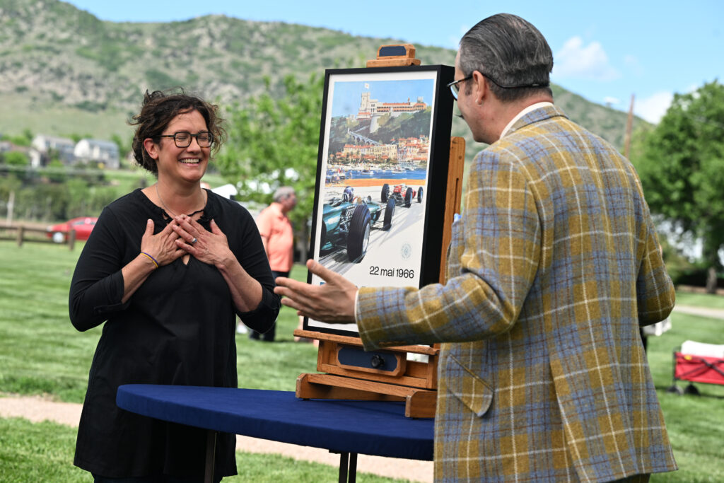 The image shows two people outdoors. The setting is a grassy park-like area with green hills and houses in the background under a blue sky with scattered clouds. The woman on the left is smiling widely and appears to be pleasantly surprised or touched. She has curly brown hair, glasses, and is wearing a black top. She’s holding her hands to her chest in a gesture that suggests gratitude or amazement. The man on the right, seen from behind and slightly in profile, is wearing a yellow and blue plaid blazer and has neatly combed gray hair. He appears to be speaking or gesturing toward the woman. Between them on a small table is an easel holding a framed vintage racing poster. The poster depicts a Formula 1 race with two cars speeding by a harbor town, likely Monaco, with the date "22 mai 1966" printed at the bottom.