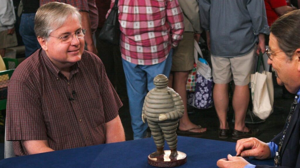 This photo shows two men seated at a blue-covered table. The man on the left is smiling slightly, wearing glasses and a short-sleeved, checkered shirt. The man on the right, partly visible in profile, is also wearing glasses and has a mustache and long hair. Between them on the table is a small, unusual figurine. The figure is shaped like a rounded, segmented human form made of stacked rings, resembling the Michelin Man (the mascot of the Michelin tire company). It's standing on a circular base with the words “MICHELIN TIRES” inscribed on it. In the background, a crowd of people is standing and milling about, many carrying bags, suggesting the setting is informal and busy. The lighting is bright and the atmosphere seems lively and social.