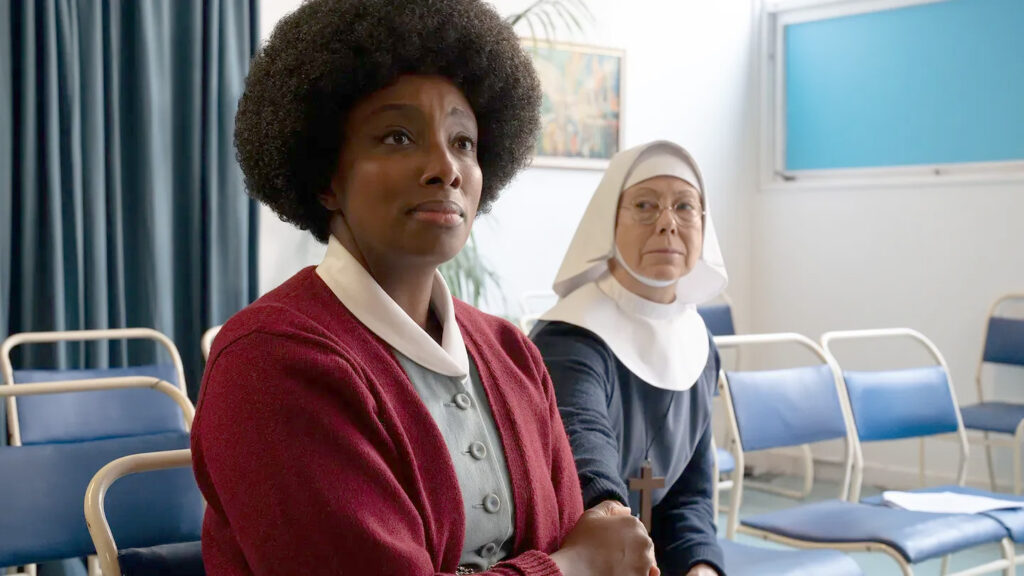 Two women sit in a waiting room with blue chairs. The woman in the foreground looks expectantly at someone off-camera, the woman behind her is looking at her and wearing a nun's garb.