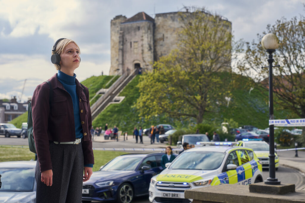 Patience stands at the left side wearing headphones and looking towards what may be a building (off screen). It is daytime and off in the distance a castle like building made of light washed bricks and a small area of grass, trees, and a lamp post.