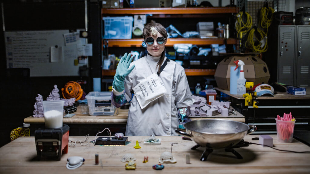 A woman dressed as a scientist stands behind a table that holds scientific equipment. Behind is another table and shelf holding more items.