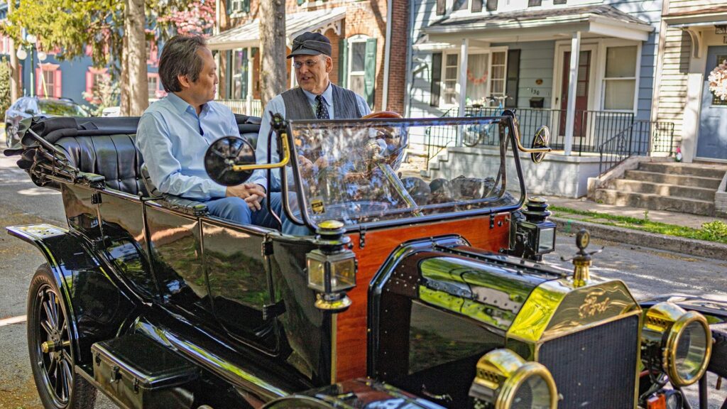 Two men sit in a vintage Ford Model T on a residential street.