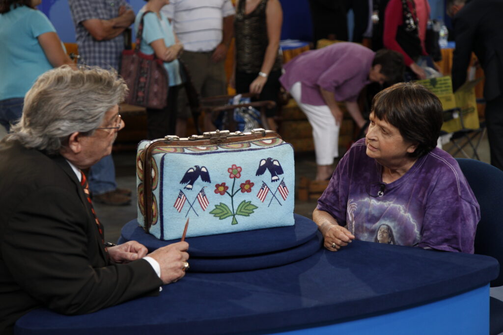A woman and a man sit at a display table that holds a vintage embroidered bag.