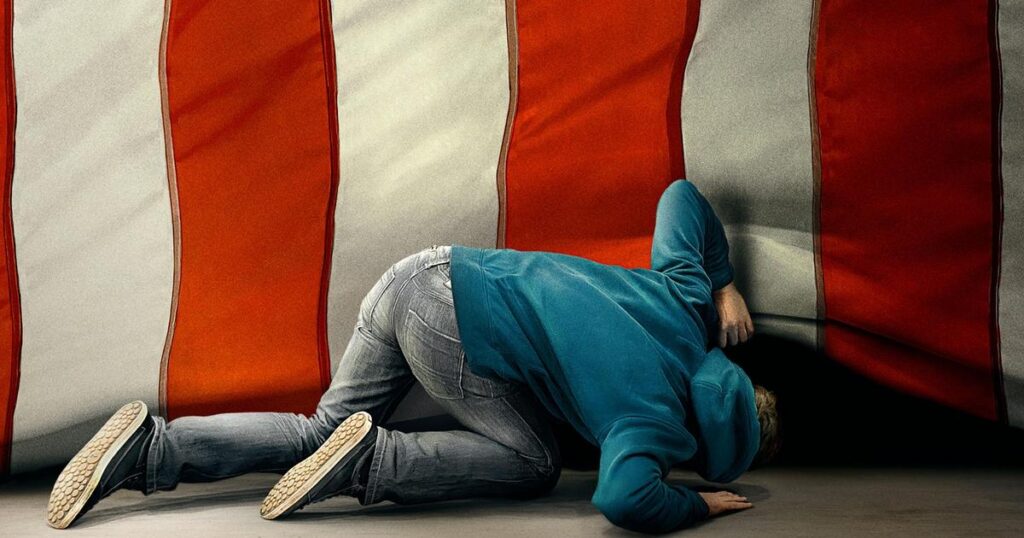 A person crouches on the floor and lifts up the bottom of a large, American Flag curtain, peering underneath it.