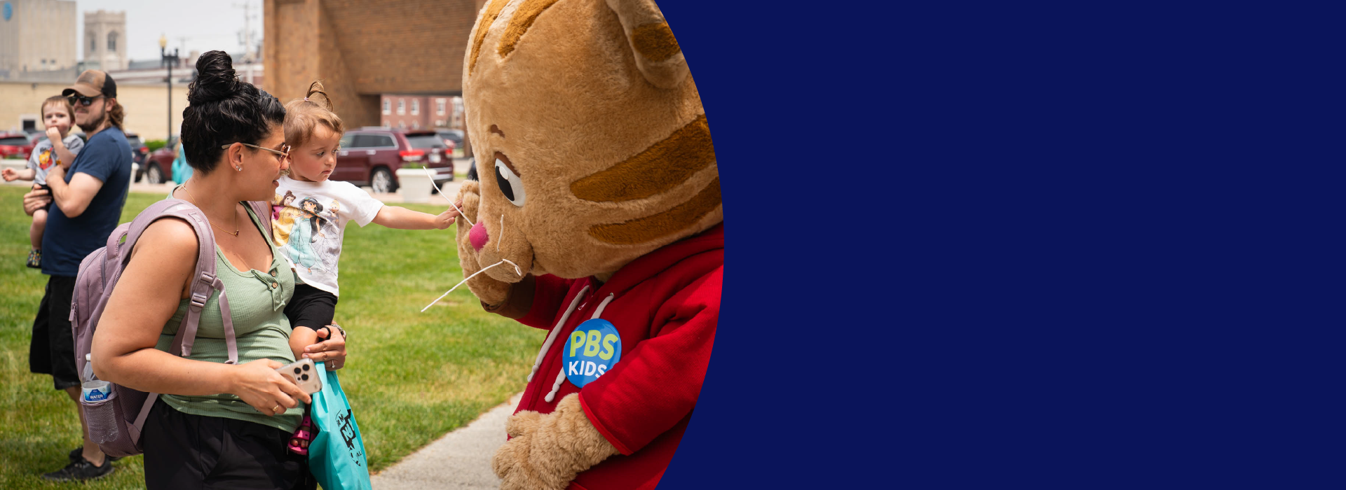 A mother holds a young child who extends her hand out to a Daniel Tiger character. Behind them is man holding another young child.
