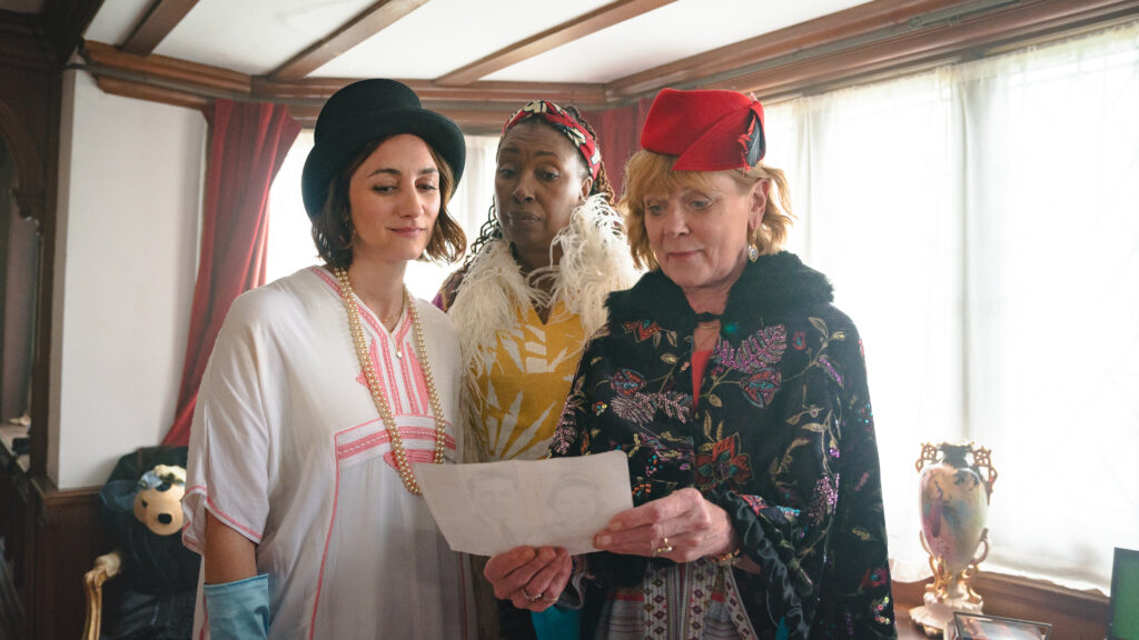 Three women dressed in eclectic, vintage outfits and distinct hats stand close together inside a wood-trimmed room, intently looking down at a piece of paper held by the woman on the right.