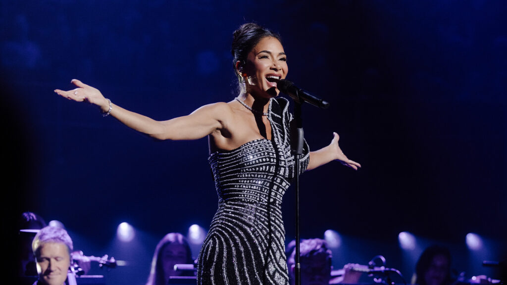 Nicole Scherzinger raises her arms in celebration as she stands on a blue-lit stage, singing into a microphone.