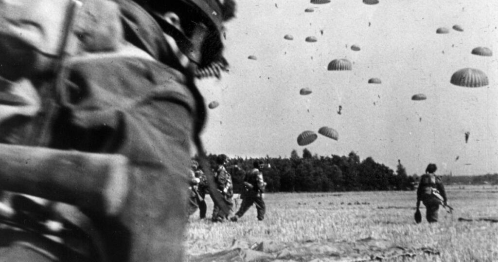 In this black and white photo, soldiers cross a large grassy field as paratroopers land around them.