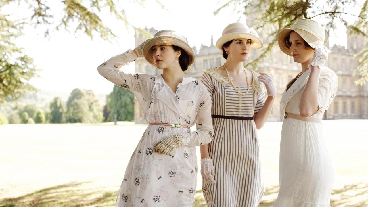 Three high-society women wearing white or light-colored dresses, gloves and hats from the early 1900s stand outside with tree branches in the foreground and a castle in the background.