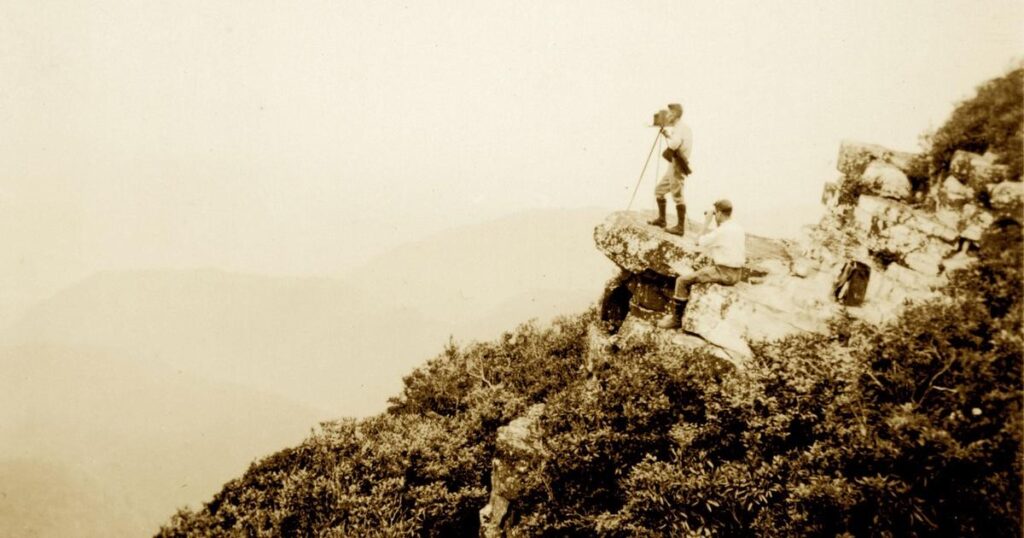 This black and white photo shows two men on a rock at the edge of a forested mountain. One sits and looks through a pair of binoculars while the other uses a tripod camera to capture the vast mountain valley.