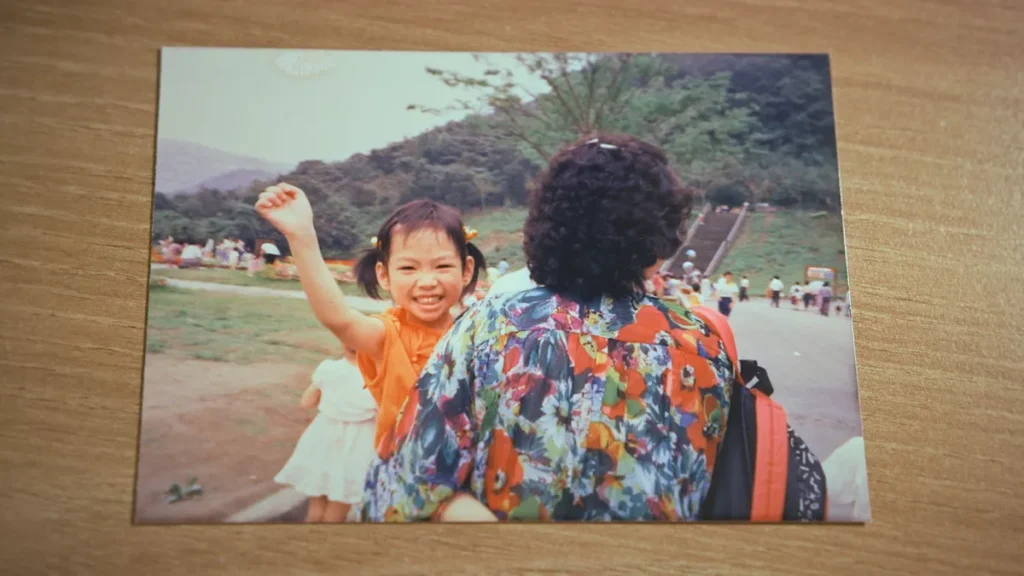 This is a picture of a printed photo. In the photo, a young Chinese girl waves at the camera. She is held by a woman who is faced away from the camera.