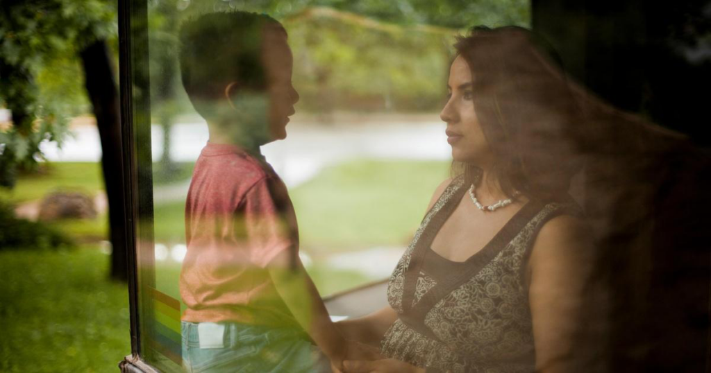 A woman and a young boy sit indoors facing each other and holding hands, as seen through a window reflecting the outdoor greenery.