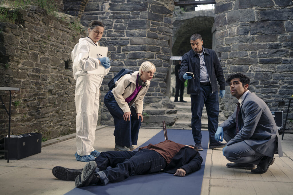 A group of investigators and a forensics expert gather around a body lying on a blue mat inside what appears to be a stone ruin or courtyard.