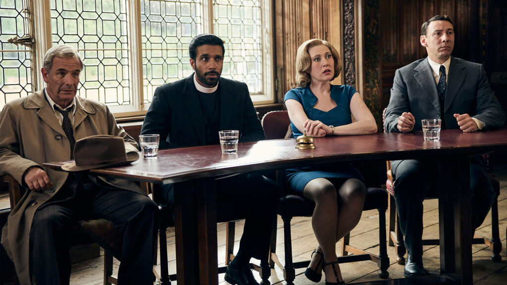 Three men in suits and a woman in a turquoise dress sit on the same side of a long wooden table. They all stare in surprise at something in the distance.
