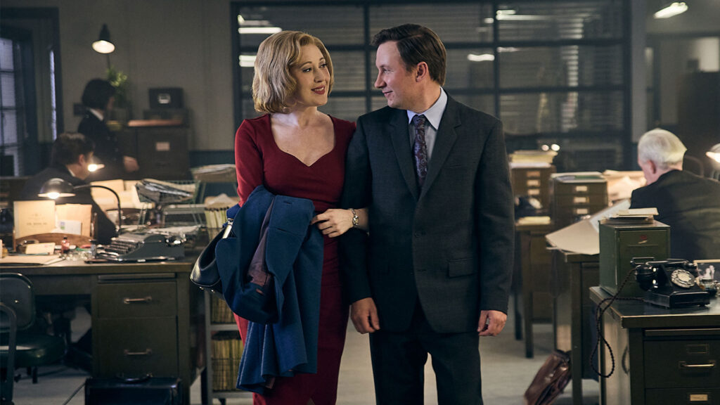 A man in a suit and a woman in a long red dress stand in the middle of an office room and look lovingly at each other. The woman holds his arm with her left hand.