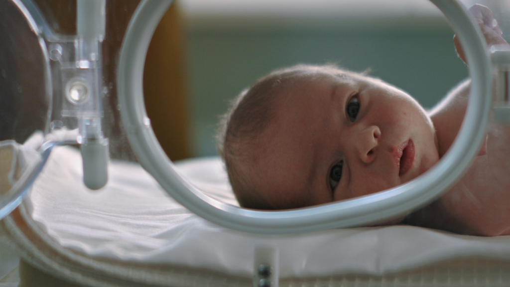 A newborn baby lays in a hospital bed, staring at the camera with wide open eyes.