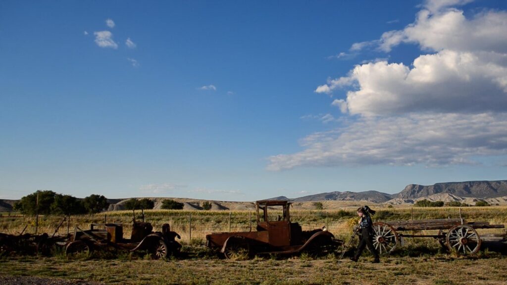 A person carrying a tripod walks past rusted, vintage vehicles that rest nearby an old wooden fence. In the background, a cloudy blue sky settles above a vast, American Southwest prairie.