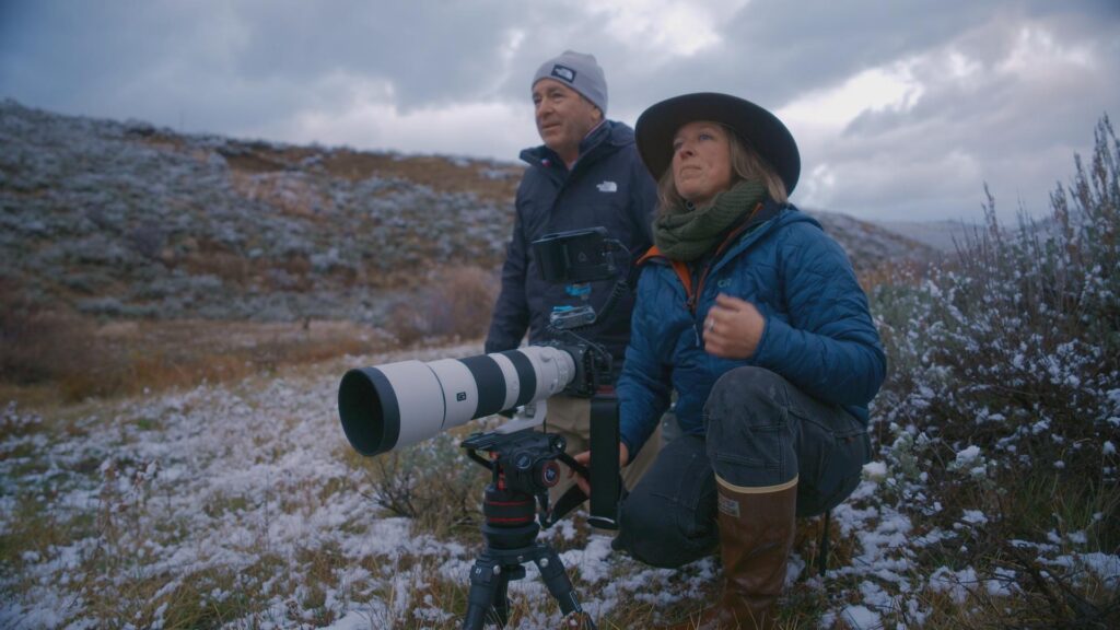 A man and woman in winter clothing crouch low to the snow covered ground. They are next to a long-distance video camera and are looking at something in the distance.