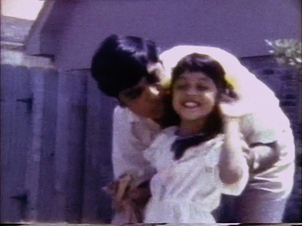 In this grainy, old photograph, a young girl smiles and waves at the camera while a man leans over and hugs her.