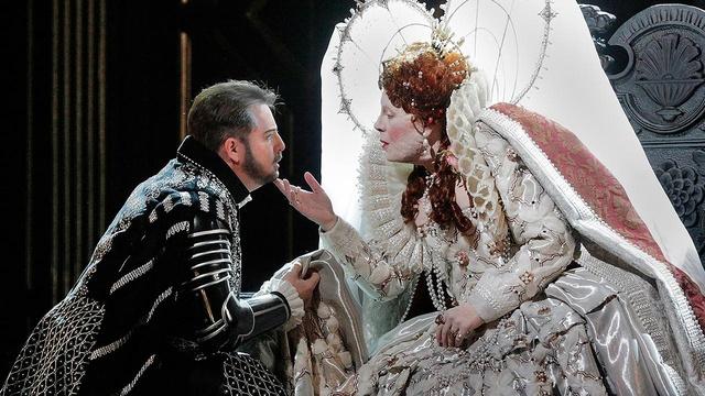 Two people in 1800's Russian costumes act dramatically on stage in this close up shot. A woman in an extravagant dress leans over the arm of her throne and touches the chin of a kneeling man.