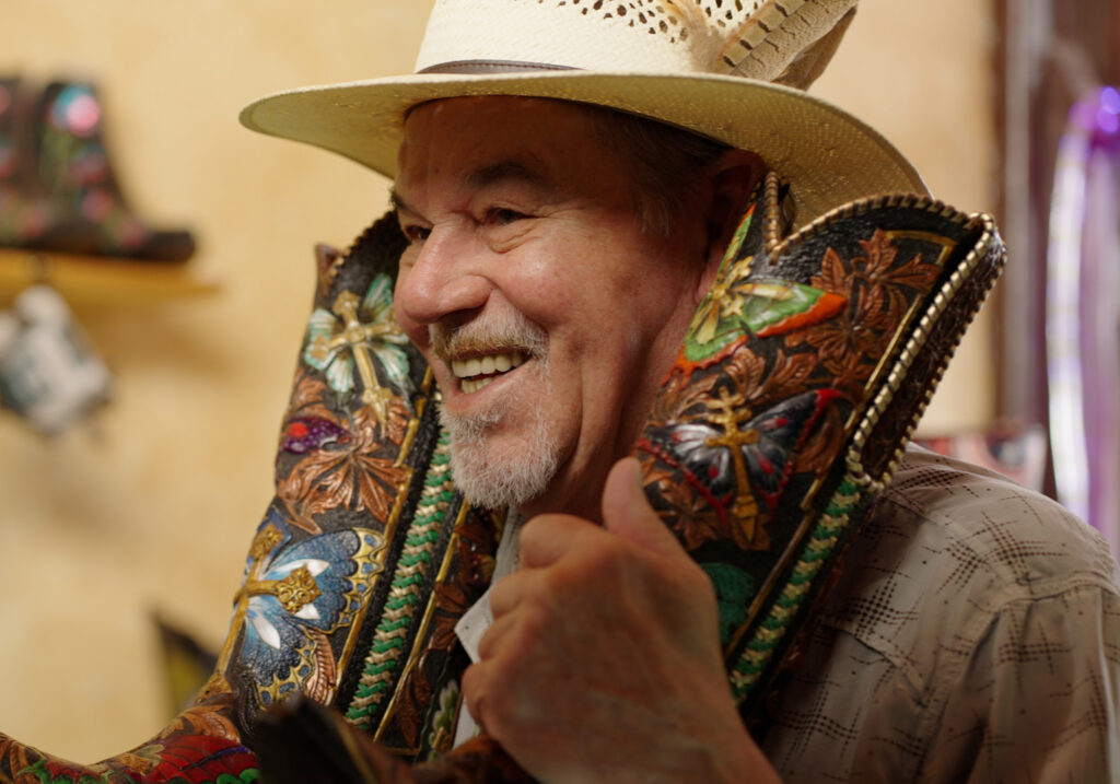 A man with a gray goatee wearing a straw cowboy hat smiles while holding a pair of colorful, decorative cowboy boots, one on each shoulder.