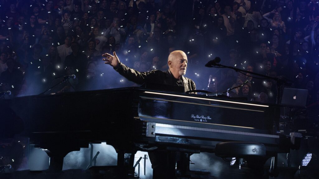 A bald man on stage behind a black grand piano and microphone