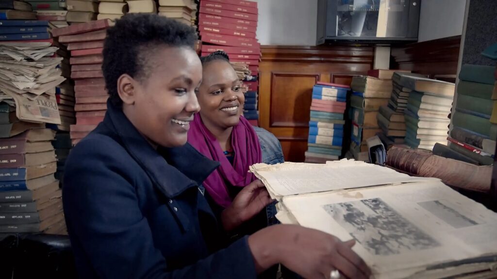 Two women look at an old book open on a table in front of them, with stacks of old books behind them.