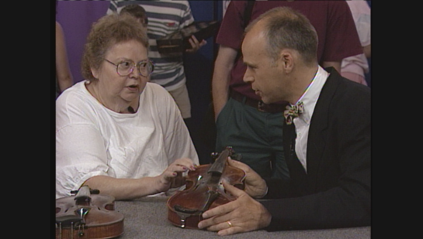 A man in a suit converses with an elderly lady while gently holding up a violin for her to touch.