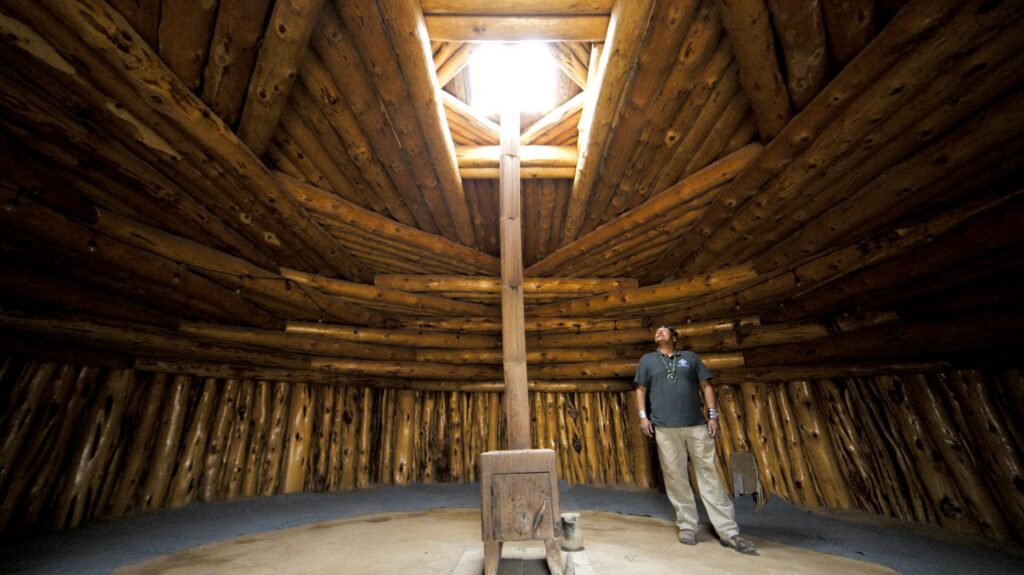 A man stands inside a round structure made of wood beam with a skylight in the middle and a stove with a chimney pipe that goes up through the skylight.