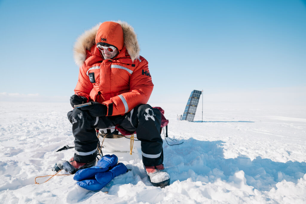 A man wears bulky, professional winter gear and crouches on a vast, snowy plain. He writes on a notepad.