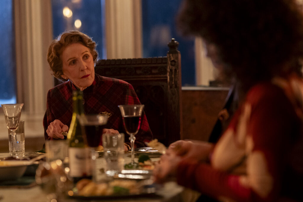 Two women sit at a dining table in a warmly lit room. One, in the foreground, is blurred and facing away, while the other leans in, intently listening.