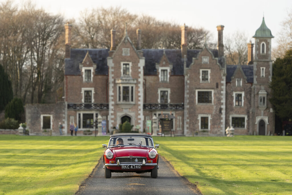 A vintage red car travels down the driveway of an old, brick mansion.