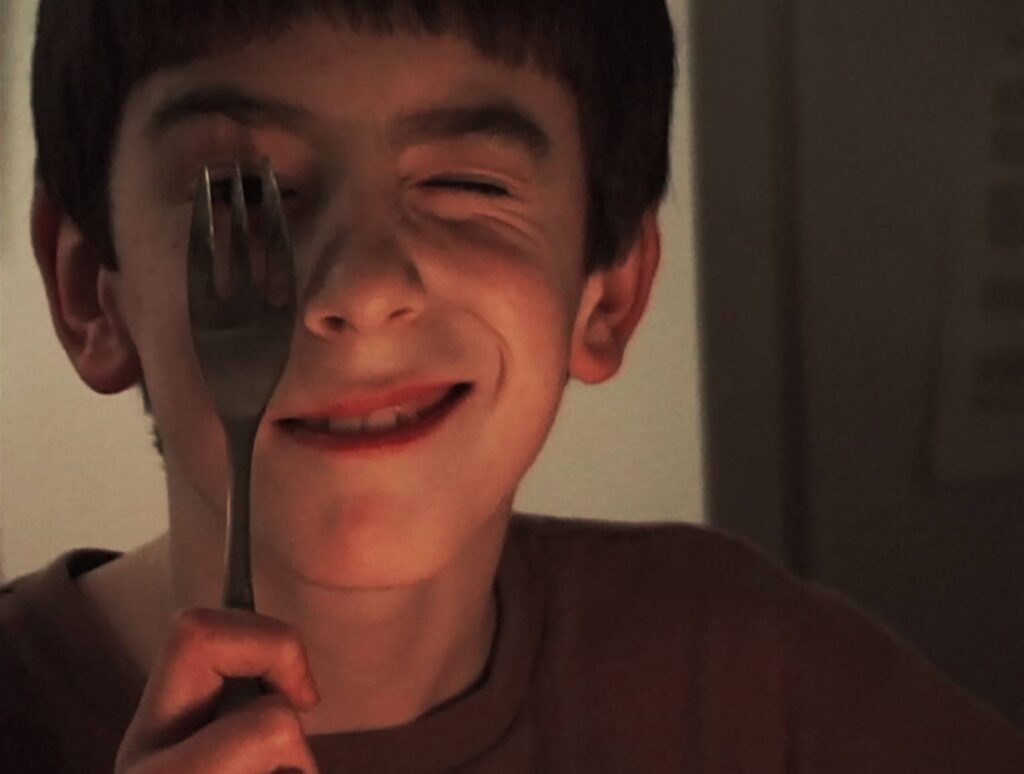 A young boy holds a metal fork up to his right eye while smiling and squinting his other eye.