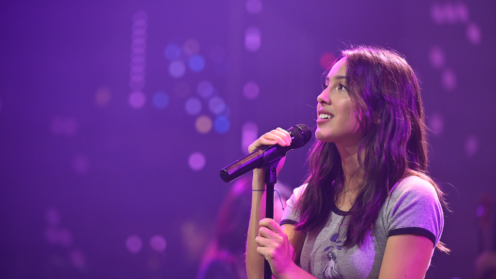Popstar Olivia Rodrigo stands on a stage that glows purple and smiles into a microphone.