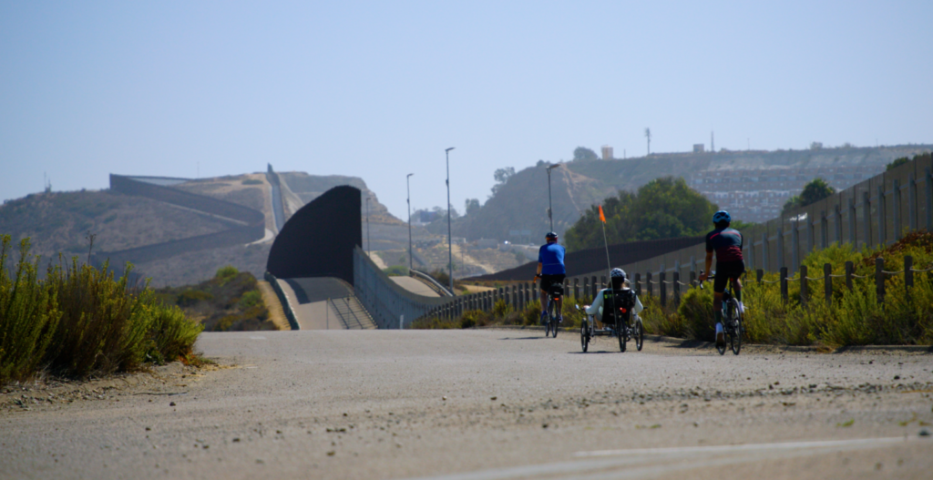 Three people ride bicycles on a path alongside a tall fence that forms a long, winding wall that stretches far into the distance.