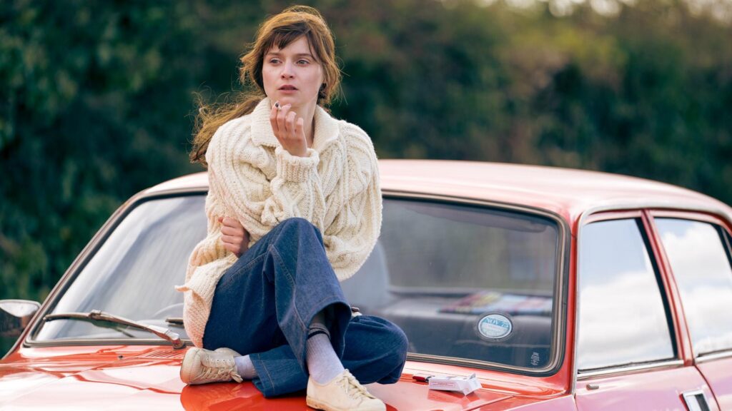 A brunette woman wearing a chunky knit sweater sits on the hood of a red car and smokes a cigarette while looking wistfully into the distance.