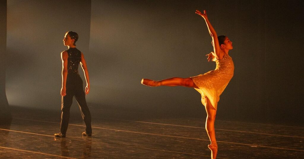 A female ballerina shines golden in the stage-light as she does a pirouette. Another ballerina stand in the background and poses strong.