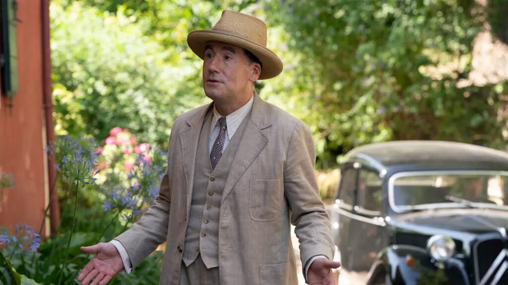 A man in a tan three-piece suit and straw hat with a vintage car behind him.