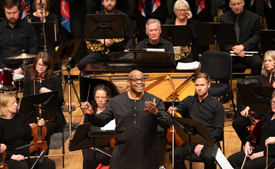 A section of an orchestra wearing all black with a man wearing glasses in the front-middle conducting